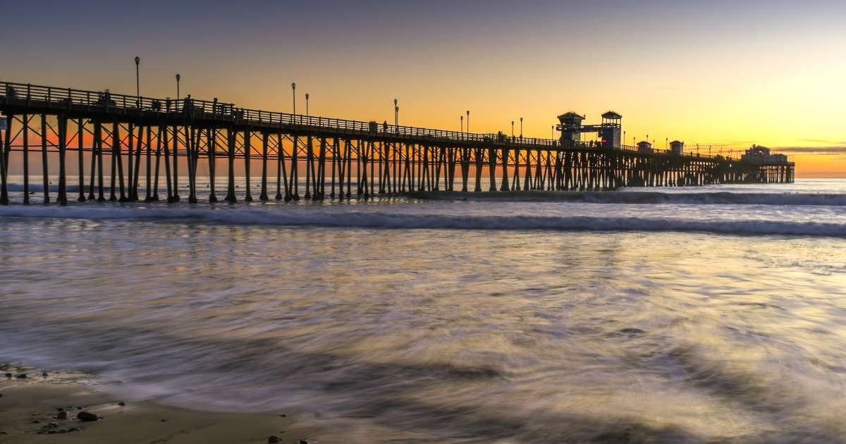 The wooden pier juts into the waters of the Pacific Ocean near a beach in Oceanside, north San Diego County, southern California, with the orange glow and purple blue sky of a dramatic sunset, in this seaside community popular with locals and tourists.