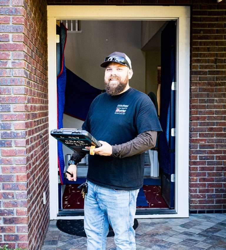 Friendly Grace Moving coordinator in gray cap holding digital tablet standing at brick home entrance for senior moving services